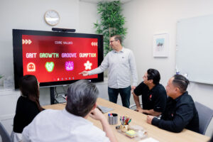 A group of people sit around a table looking at a man give a presentation
